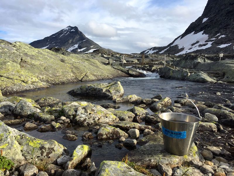 Image of river coming out of lake after a wooden bridge. A bucket used to collect water for drinking is shown. The picture is taken in Norway's Jotunheimen National Park by the Olavsbu hut.